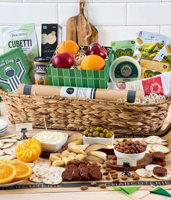 The image features a large basket filled with fresh fruits, cheese, crackers, olives, and snacks. Game day-themed decorations, such as a foam finger and mini football field, enhance the festive presentation. 