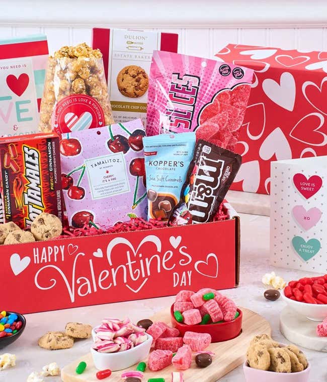 Valentine's Day gift box with heart designs, filled with kettle corn, gummies, chocolates, wafer cookies, and candies, displayed alongside bowls and plates of treats.