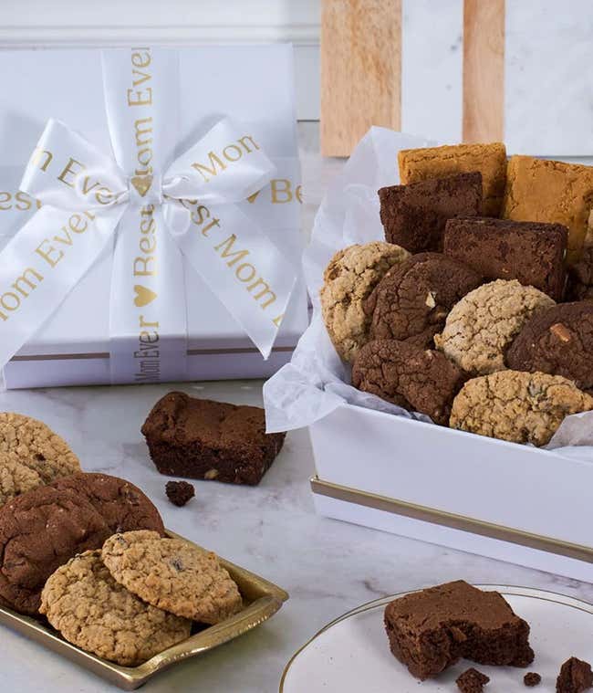 Gift box filled with assorted cookies and brownies, including chocolate chip, oatmeal, and fudge varieties, displayed on a marble surface with a white and gold Best Mom Ever ribbon tied around the box.