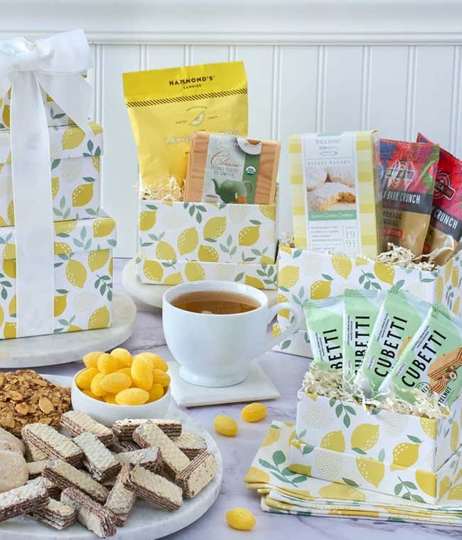 Lemon-themed gift tower with yellow and white boxes filled with wafers, cookies, lemon candies, snack bars, crackers, and tea, displayed around a cup of tea on a marble surface.