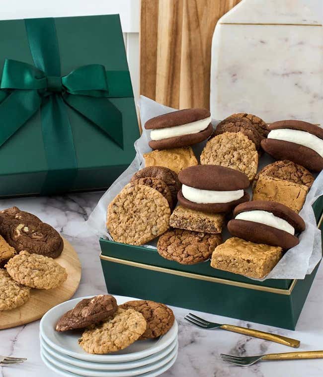 Green gift box with a satin bow filled with assorted baked treats including oatmeal cookies, chocolate chip cookies, blondies, brownies, and whoopie pies, displayed on plates and cutting boards.