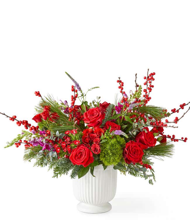 Festive holiday bouquet with red roses, red spray roses, burgundy snapdragons, and burgundy sweet william arranged with pine greenery and red berry accents in a white ribbed ceramic vase.