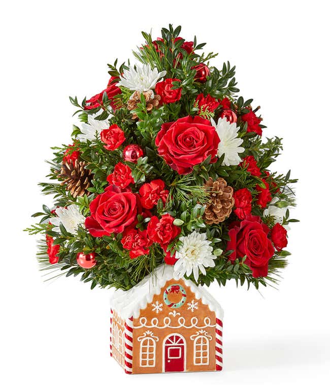 Mini Christmas tree arrangement featuring red roses, miniature red carnations, and white cushion poms accented with festive greenery, pinecones, and red ornaments. Displayed in a charming gingerbread house container.