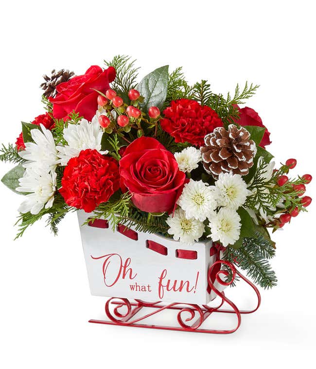 Festive holiday bouquet featuring red roses, red carnations, white button poms, and white cushion poms accented with lush seasonal greenery. Arranged in a cheerful container, this classic red and white floral display captures the joyful spirit of the seas