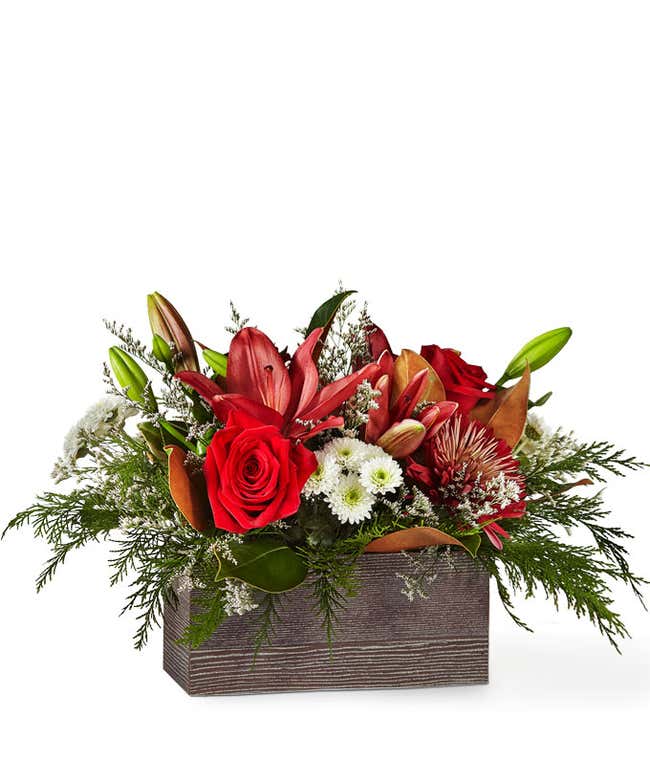Holiday floral arrangement featuring red roses, lilies, white poms, and greenery artistically arranged in a rustic wooden container.