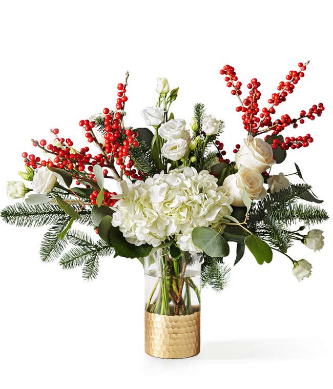 Festive winter bouquet featuring white hydrangeas, roses, and lisianthus accented with bright red berry branches, lush greenery, and pine sprigs arranged in a clear vase with a hammered gold base.