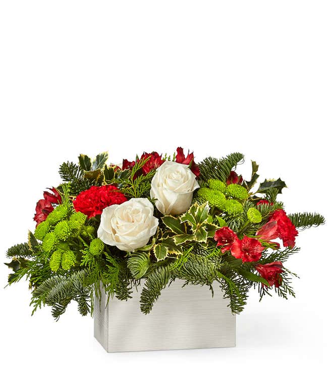 Festive floral arrangement featuring white roses, red carnations, green button poms, holly leaves, and evergreen branches, arranged in a modern white rectangular container.