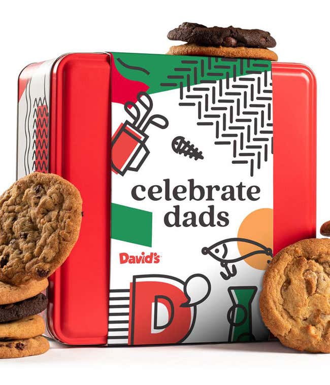 A red tin from David's with a celebrate dads-themed design, featuring illustrations of tools and a fishing rod. Assorted cookies, including chocolate chip and double chocolate, are stacked on top of and around the tin.