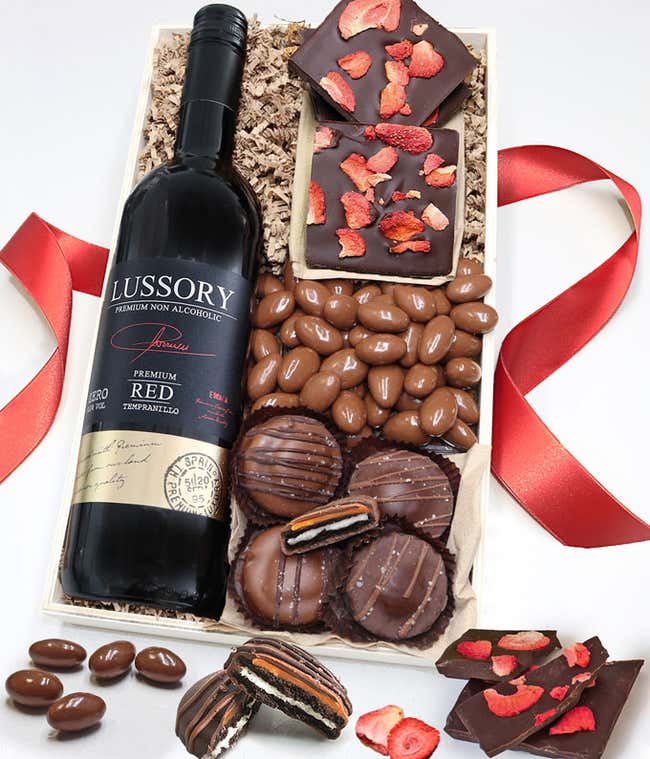 A gift box of chocolate covered Oreos topped with caramel, Dark Chocolate bark topped with dried strawberries, &amp; a bag of chocolate covered almonds next to a bottle of non-alcoholic red wine