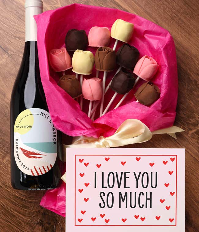 A bouquet of Belgian chocolate roses in milk, dark, and white chocolate, wrapped in pink tissue paper and tied with a cream ribbon, accompanied by a bottle of red wine and card reading 