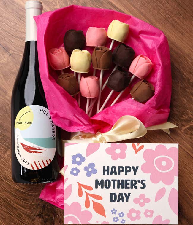 A bouquet of Belgian chocolate roses in milk, dark, and white chocolate, wrapped in pink tissue paper and tied with a cream ribbon, accompanied by a bottle of red wine and card reading 