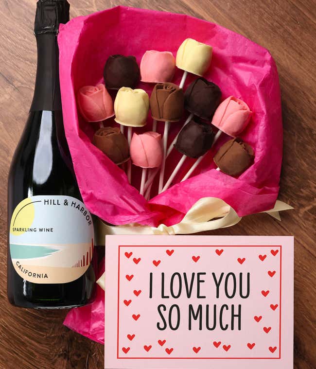 A bouquet of Belgian chocolate roses in milk, dark, and white chocolate, wrapped in pink tissue paper and tied with a cream ribbon, accompanied by a bottle of sparkling wine and card reading 