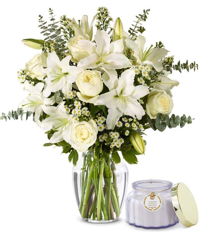 An elegant floral arrangement featuring white roses, white lilies, white asters, and eucalyptus, optionally displayed in a glass vase. Sitting next to a purple candle
