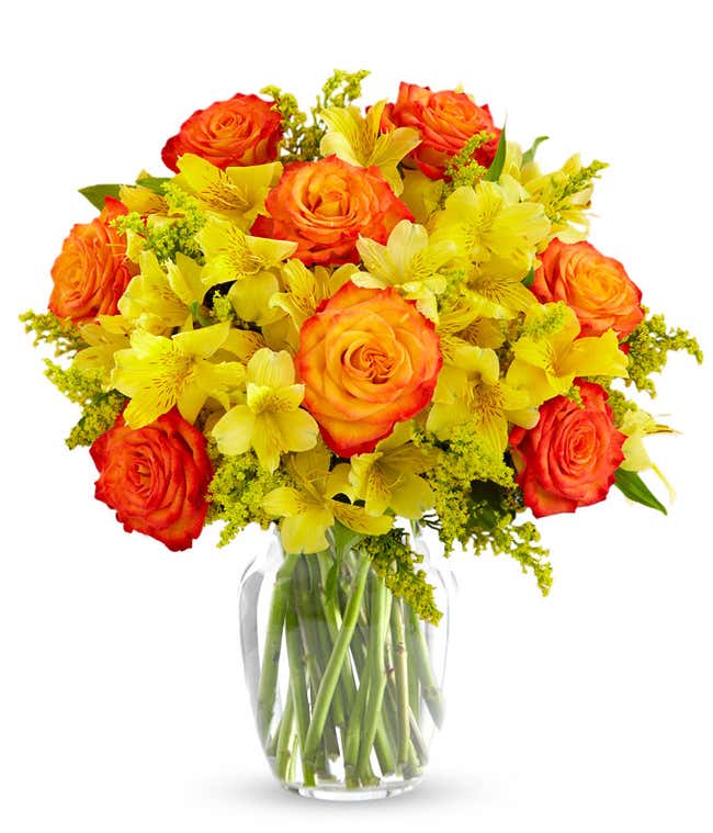 A bouquet of yellow alstroemeria, orange roses with lighter orange centers, yellow solidago in a clear vase on a white background.