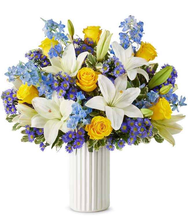 An elegant floral arrangement featuring white Asiatic lilies, yellow roses, blue delphinium, and purple Monte Casino, optionally arranged in a glass vase. This vibrant mix offers a stunning and colorful display.