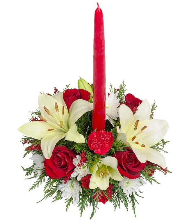 Festive centerpiece featuring a tall red candle surrounded by white lilies, red roses, red carnations, white poms, and greenery.