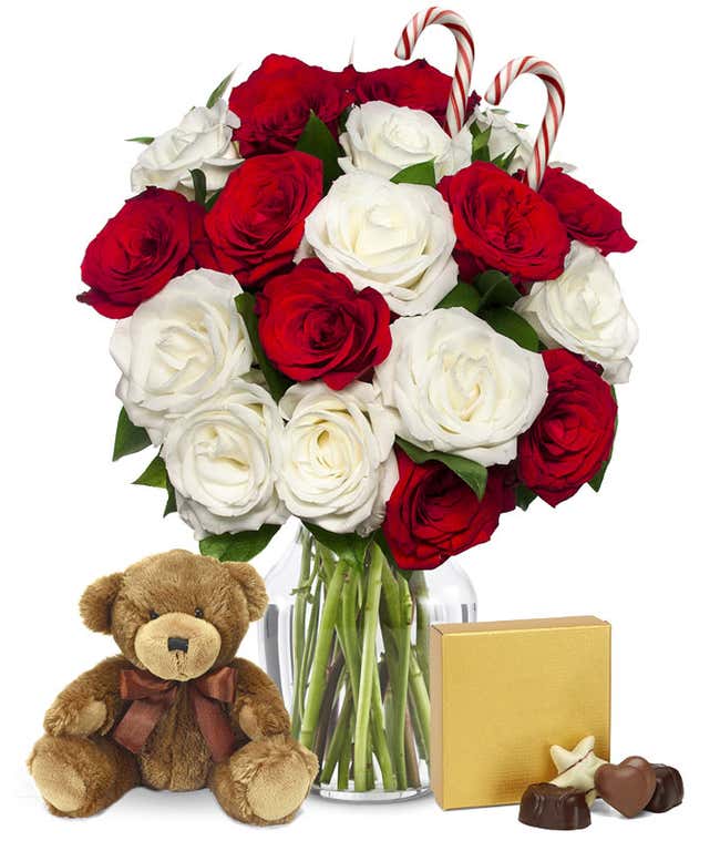A bouquet of red and white roses in a clear vase, accented with two candy canes. In front of the vase are a brown teddy bear with a ribbon and a gold gift box with a bow, creating a festive holiday display.