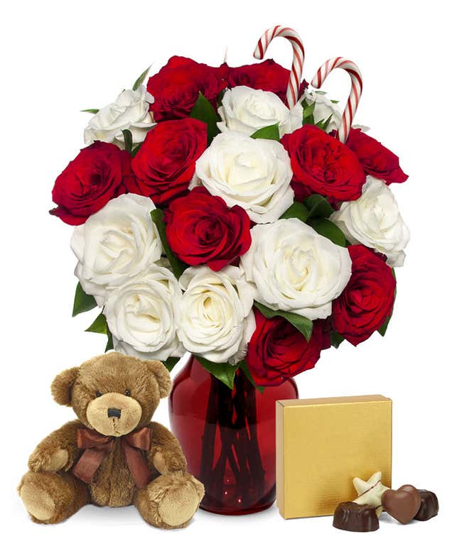 A bouquet of red and white roses in a clear vase, accented with two candy canes. In front of the vase are a brown teddy bear with a ribbon and a gold gift box with a bow, creating a festive holiday display.