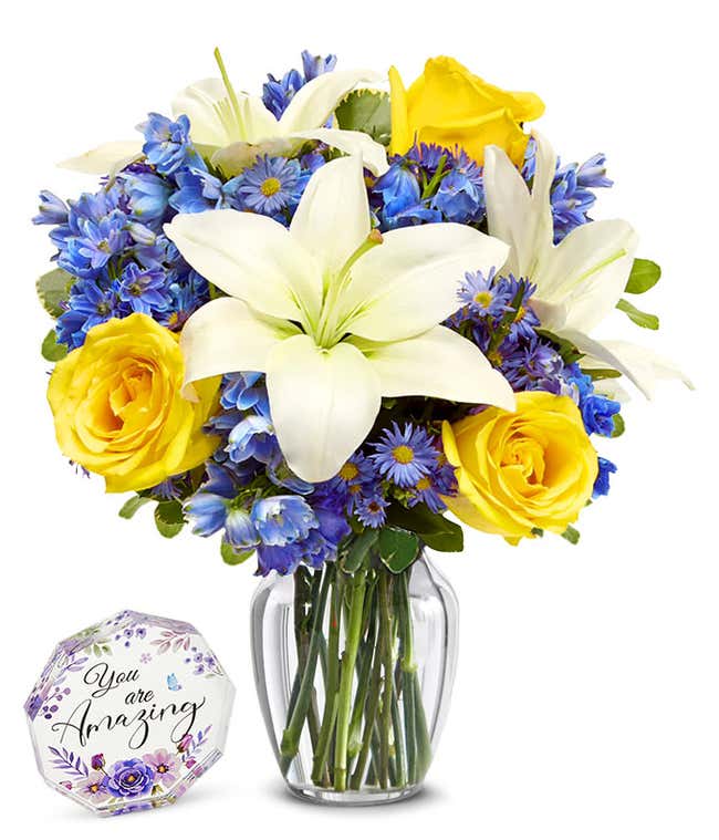 An elegant floral arrangement featuring white Asiatic lilies, yellow roses, blue delphinium, and purple Monte Casino. Next to a nonagon clear acrylic keepsake reading 'you're amazing' with purple flowers.