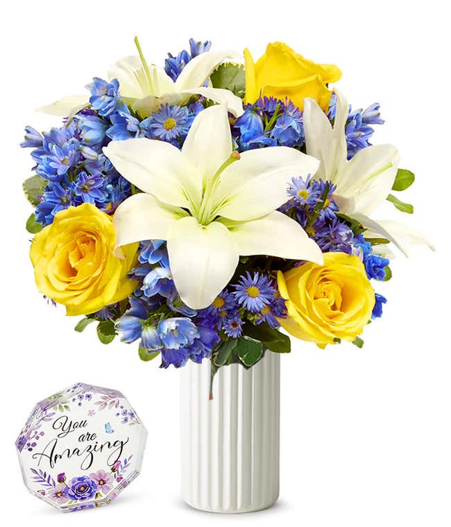 An elegant floral arrangement featuring white Asiatic lilies, yellow roses, blue delphinium, and purple Monte Casino. Next to a nonagon clear acrylic keepsake reading 'you're amazing' with purple flowers.