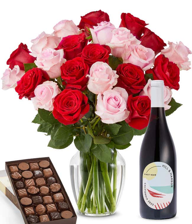 A bouquet of 24 red and pink roses arranged in a glass vase is displayed beside a bottle of Hill &amp; Harbor Pinot Noir and a 1 pound box of assorted chocolates.