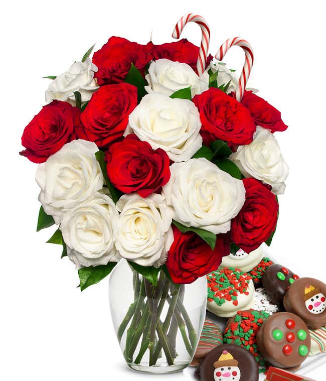 9 red and 9 white roses and 2 candy canes popping out of a glass vase with some leaves showing. Next to it is a plate of chocolate cookies with red/green sprinkles, candy, and snowmen.