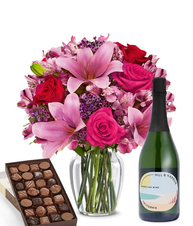 A mixed bouquet of pink and purple flowers in a clear glass vase is paired with a bottle of Hill &amp; Harbor Sparkling Wine and a box of assorted chocolates.