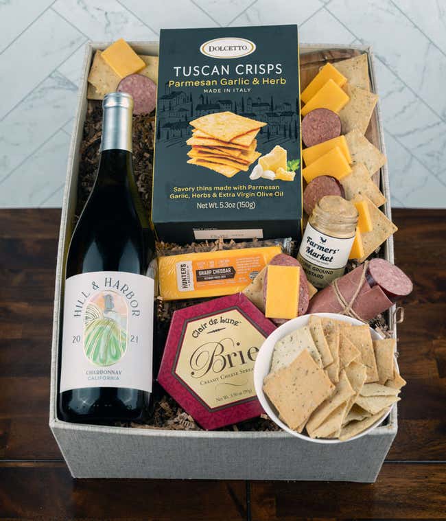 a gift basket with contents laid out across a white wood table. A bottle of white wine, a glass of white wine, a block of cheddar cheese, a summer sausage, a jar of honey mustard, a box of crackers, with a cutting board of sliced meat and cheese 