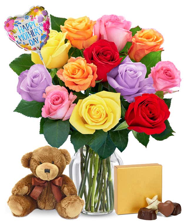 One Dozen Rainbow Roses in a clear glass vase with a box of chocolates, a teddy bear, and a heart shaped Happy Mother's Day mylar balloon