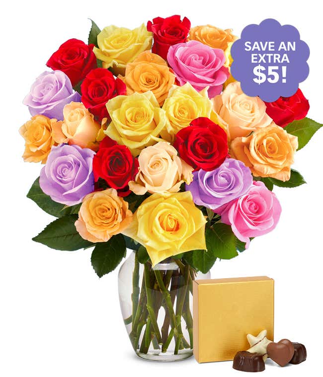 Mixed bouquet of vibrant roses in red, yellow, pink, orange, lavender, and cream displayed in a clear glass vase, accompanied by a gold box of assorted chocolates.