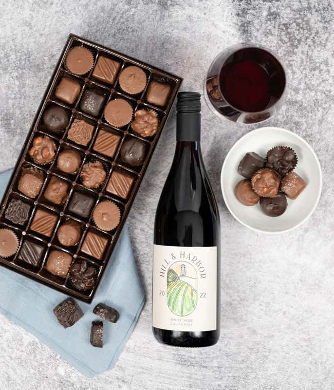 a bottle of red wine and a box of assorted chocolates laid out on a table with glasses of wine, cloth napkins, loose chocolates, and a cutting board