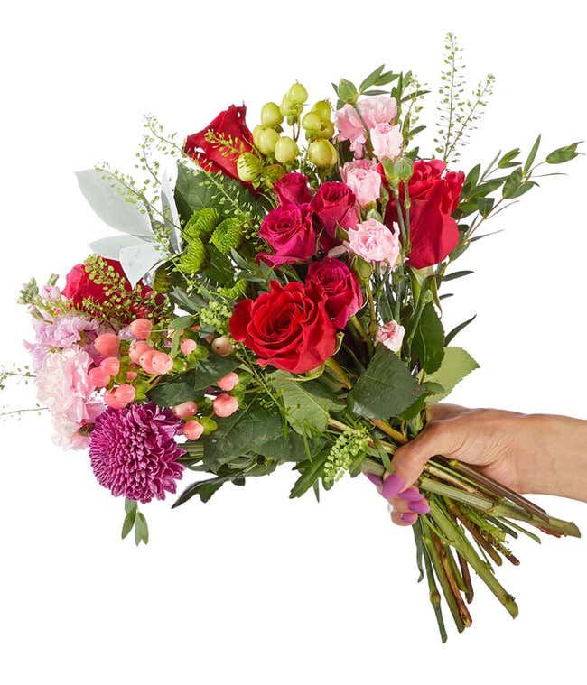 A hand-tied bouquet of red and pink roses, spray roses, light pink and hot pink spray roses, soft light pink stock and fresh greenery arranged in a lush, romantic style.