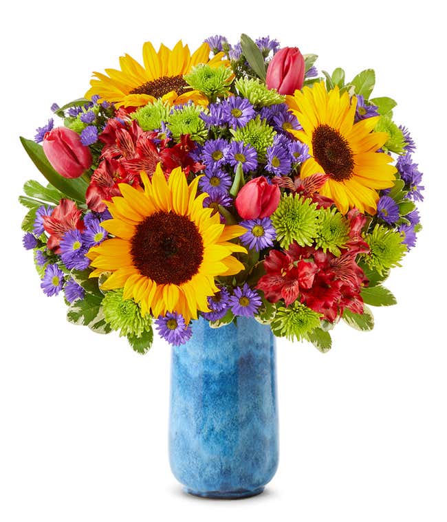 A vibrant arrangement featuring sunflowers, pink tulips, red alstroemeria, purple Monte Casino asters, and green poms, optionally arranged in a glass vase. This lively mix offers a bright and cheerful display.