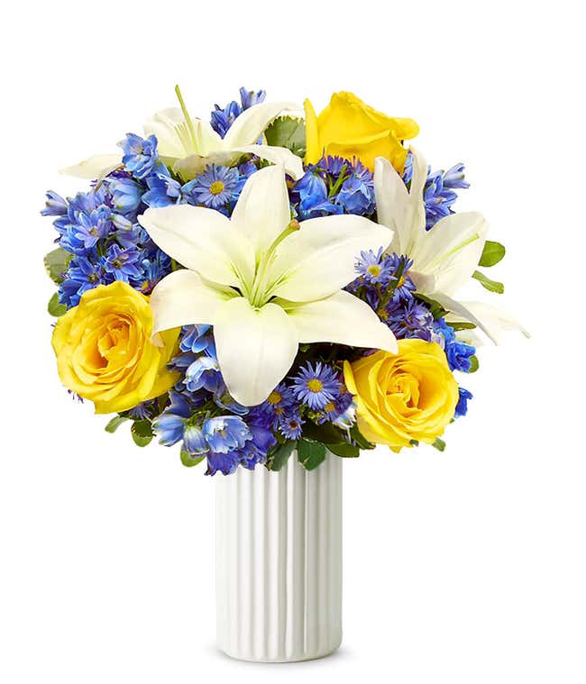 An elegant floral arrangement featuring white Asiatic lilies, yellow roses, blue delphinium, and purple Monte Casino, optionally arranged in a glass vase. This vibrant mix offers a stunning and colorful display.