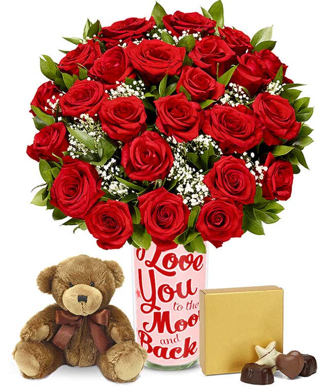 a bouquet of 24 red roses with small white flowers and greenery in between in a clear glass vase with a small gold box of chocolates and a small brown teddy bear next to it