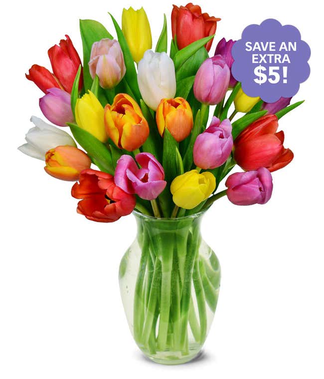 A clear glass vase filled with a vibrant bouquet of multicolored tulips, including red, yellow, orange, purple, pink, and white blooms, with fresh green leaves.