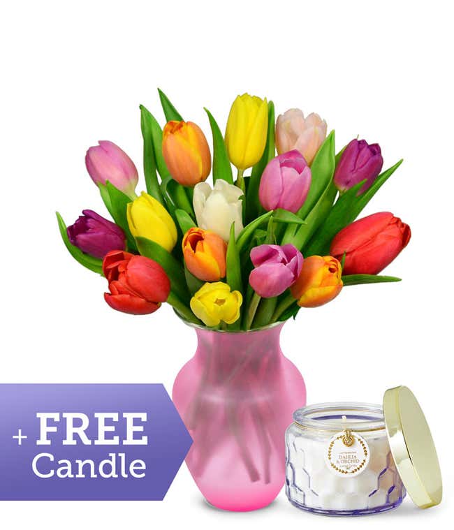 A clear glass vase filled with a cheerful bouquet of multicolor tulips in red, yellow, pink, orange, purple, and white, displayed alongside a dahlia and orchid scented candle in a clear glass jar.