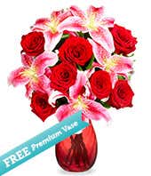 A bouquet in a glass vase of pink and red roses, pink lilies, pink and purple filler flowers sitting next to a small purple candle in glass jar. 