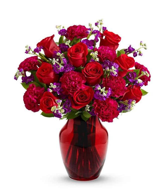 A bouquet of red roses, red carnations, and purple stock in a glass vase.
