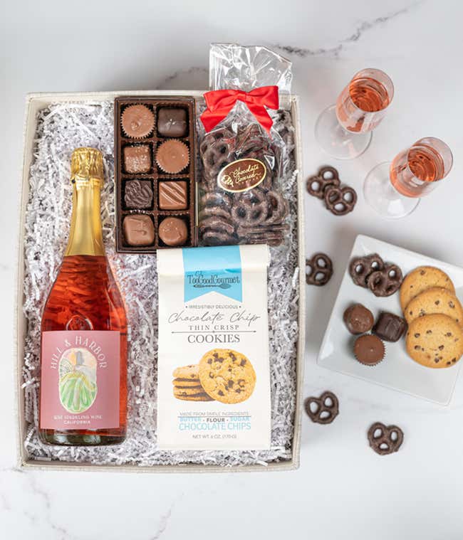 an overhead view of a gift box with choclates, chocolate pretzels, chocolate chip cookies, and a bottle of pink sparkling wine