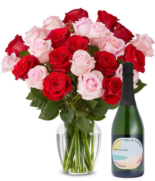 A mix bouquet of two dozen red and blush roses fills a clear glass vase and is paired with a bottle of Hill &amp; Harbor California Sparkling Wine.