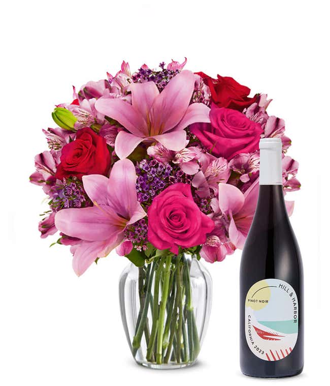 A bouquet of pink lilies, pink roses, and greenery arranged in a clear glass vase sitting next to a bottle of Hill &amp; Harbor California Pinot Noir.