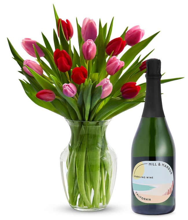 A clear glass vase filled with 15 stems of mixed red and pink tulips and bright green stems sits beside a bottle of Hill &amp; Harbor California Sparkling Wine.