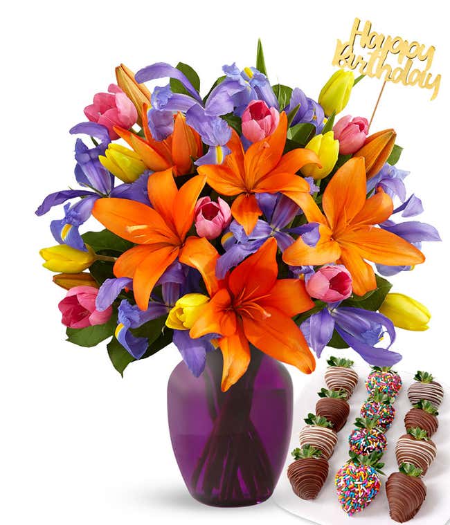 Bright bouquet of orange lilies, purple irises, and multicolored tulips in a clear glass vase with a gold Happy Birthday pick, paired with a plate of chocolate-covered strawberries featuring swizzled and rainbow sprinkle designs.