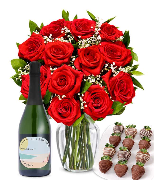 A bouquet of 1 dozen red roses in a clear glass vase is displayed beside a bottle of Hill &amp; Harbor Sparkling Wine and an arrangement of 12 chocolate-covered strawberries.