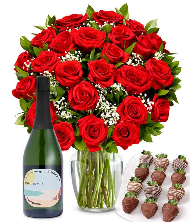 A bouquet of 2 dozen red roses in a clear glass vase is displayed beside a bottle of Hill &amp; Harbor Sparkling Wine and an arrangement of 12 chocolate-covered strawberries.