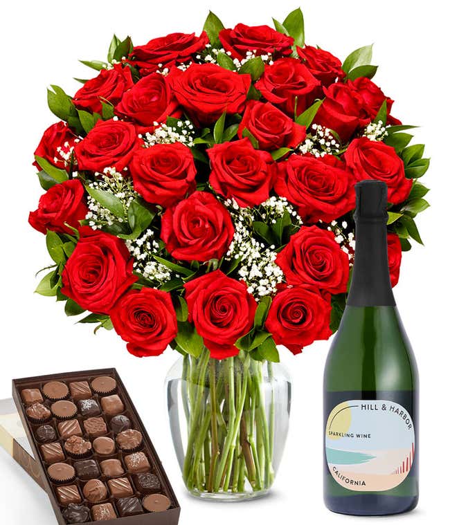 A lush arrangement of 2 dozen red roses in a clear vase sits beside a bottle of Hill &amp; Harbor Sparkling Wine and a 1-pound box of gourmet assorted chocolates.