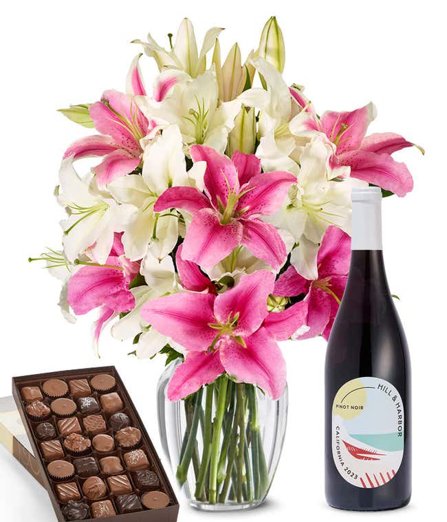 A bouquet of pink and white lilies arranged in a clear vase sits beside a bottle of Hill &amp; Harbor Pinot Noir and box of assorted chocolates.