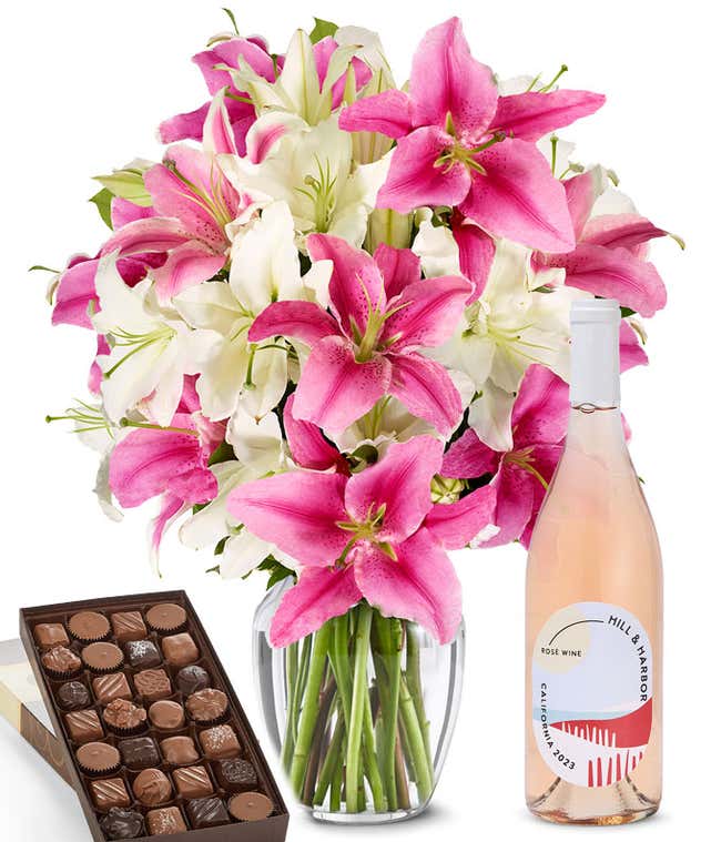 A bouquet of pink and white lilies arranged in a clear vase sits beside a bottle of Hill &amp; Harbor Ros&eacute; and box of assorted chocolates.