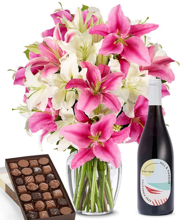 A bouquet of pink and white lilies arranged in a clear vase sits beside a bottle of Hill &amp; Harbor Pinot Noir and box of assorted chocolates.